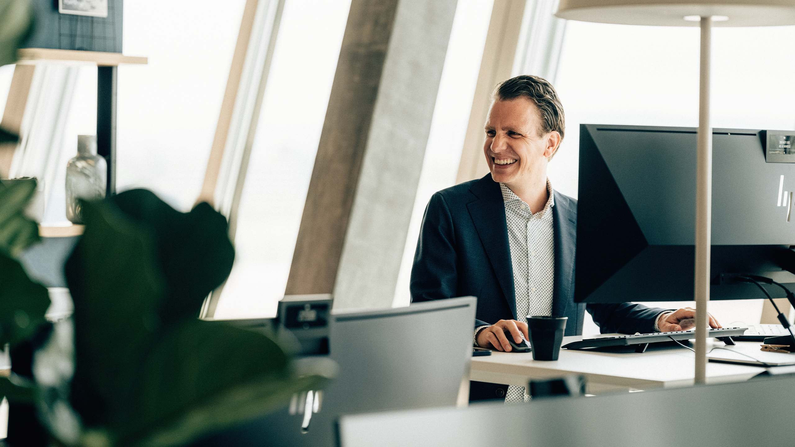 Lachender Mann vor dem Computer am Arbeitsplatz in einem offenen Büro