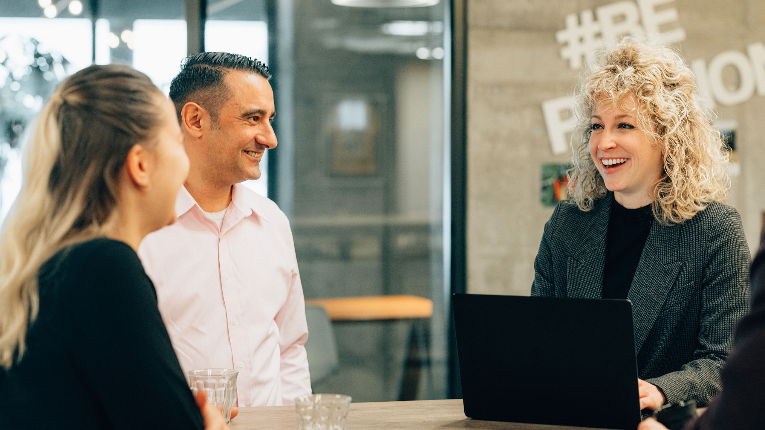 Lachende blonde Frau am Laptop spricht mit zwei anderen Personen